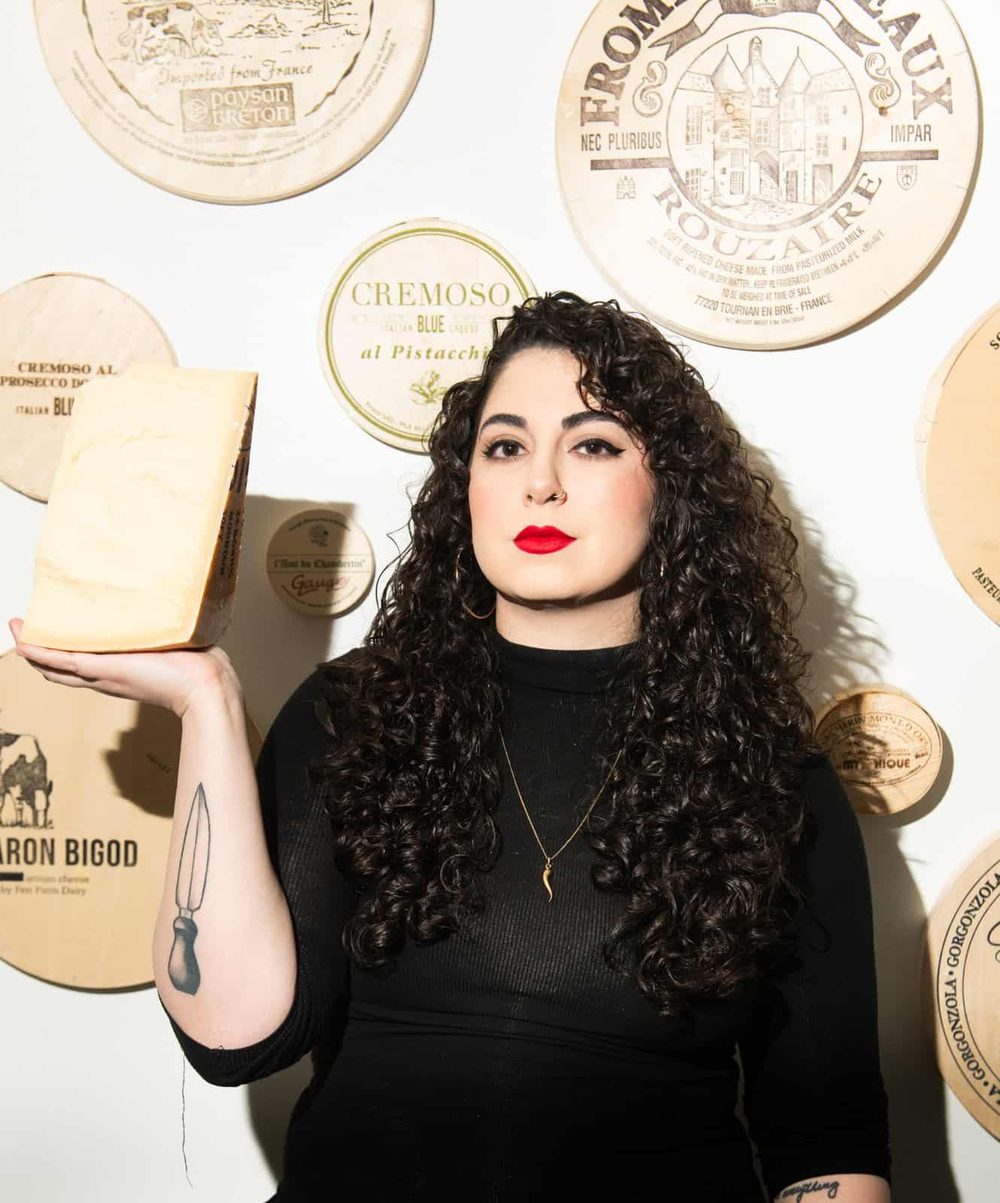 Emilia in black holding a wedge of hard cheese against a wall of cheese-box lids
