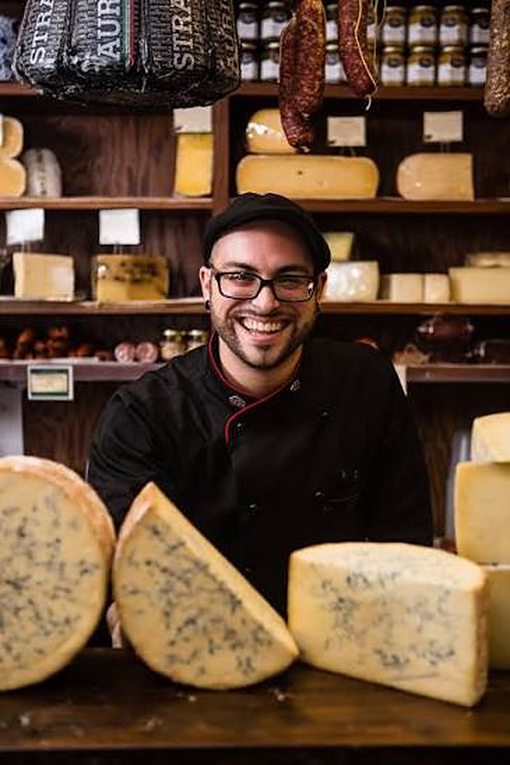 Tommy smiling behind three wedges of blue cheese in a cheese shop